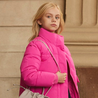 Young girl wearing a bright pink puffer jacket holding a beige handbag against a beige wall.