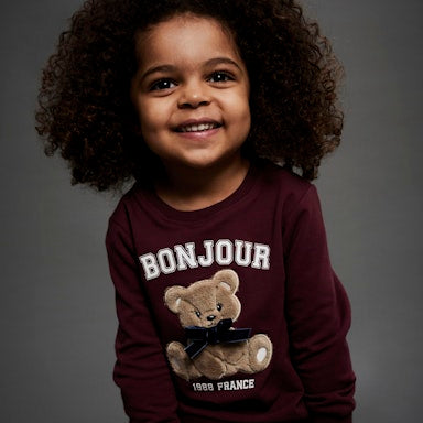 Child wearing a maroon sweatshirt with a teddy bear graphic and 'Bonjour' text on a grey background