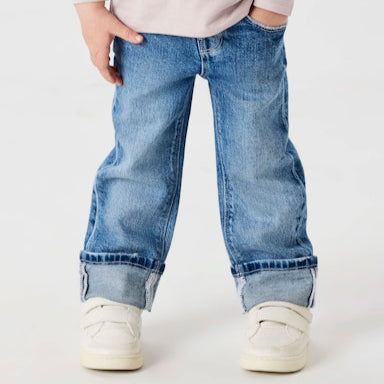 Child wearing a light pink sweatshirt with a graphic and text, blue jeans, and white sneakers on a plain background